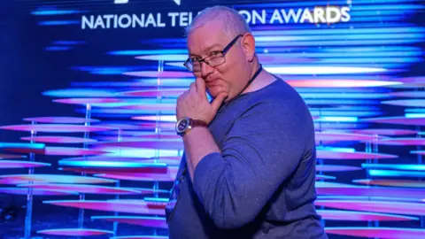'Fantasist' promised music stars for festival that never happened Rick Bronks James Kenny is standing sideways in front of a blue and purple promotional screen at the National Television Awards, with the NTA branding behind him. He is a white man with short grey, thinning hair who is smiling and holding his hand up to his chin in a comedic way. He is wearing a blue jumper, square glasses and a NTA lanyard.