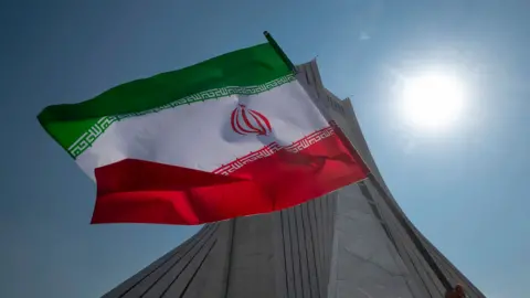 UK faces rising and unpredictable threat from Iran, report warns NurPhoto via Getty Images An Iranian flag is held in the air by a person out-of-frame in front of the Azadi monument against a blue sky and bright sun, in Tehran in February 2024