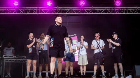 London Pride returns as events struggle with falling funds OhMyMedia / Olly Rigby Reece stands on stage at the Pink Picnic event in Salford. With pink stage lights above him, he is holding a microphone by his side and has colleagues applauding behind him. His is wearing a black polo top with Salford Pride written on the side and blackshorts.