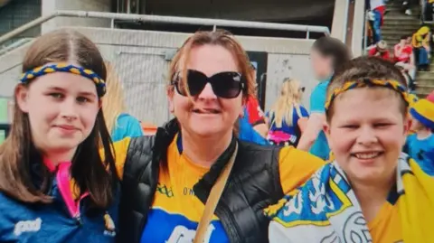 PSNI A woman standing in between a teenage girl and a teenage boy. They are wearing GAA clothes. 