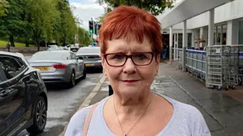 Food prices remain 'stubbornly high' in Northern Ireland as inflation rate rises A woman with short red hair is standing in the street outside a supermarket. The ground is wet. She's wearing a grey top and a silver necklace.