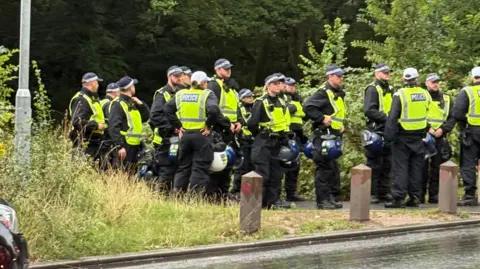 Epping council votes to urge government to shut asylum hotel Nadira Tudor/BBC A large group of police officers, more than 15, are stood on a public path near a rain soaked road, many are holding police helmets in their hands. All are wearing high-vis vests.