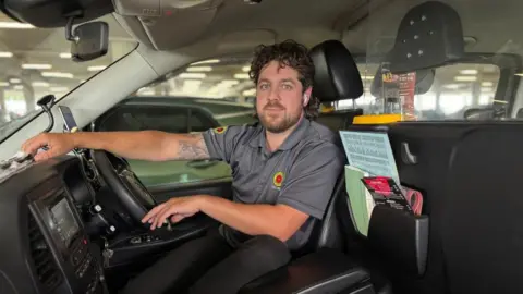 Airport drivers try to beat drop-off charges by parking in laybys Dean Carse smiling right at the camera while sitting in the cab of his taxi. He is wearing a grey polo shirt with a red and yellow logo on the upper right chest and left sleeve. He has dark curly hair and a short, dark bear. Part of a tattoo is visible on his upper right arm, which is resting on the dashboard.