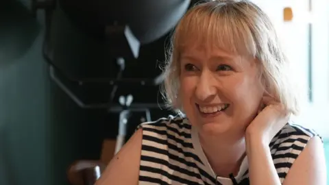 Laura Price has short blonde hair.  She is wearing a blue and white striped vest top.