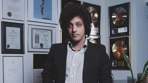 Billy Joel feels 'good' after brain disorder diagnosis Michael Putland/Getty Images A man is looking directly at the camera, with a lot of frizzy dark hair. He is wearing a blue striped button down shirt with a black jacked, and he's standing in front of a wall of framed awards.