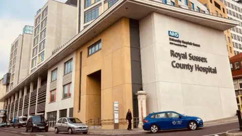 Surgeon banned by private practice is working for NHS The exterior of the Royal Sussex County Hospital in Brighton, with the name of the hospital written on the side of the building. A pedestrian is walking into the entrance, being passed by a blue taxi. Several other vehicles are parked on the side of the world.