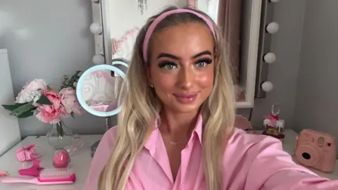 HANDOUT A young woman with long blonde hair wearing a pink shirt taking a selfie with beauty accessories in the background 