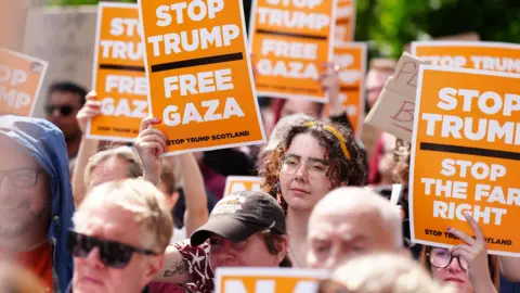 Security tightens as Donald Trump plays golf at Turnberry PA Media A crowd of anti-Trump protesters with orange signs bearing slogans including "Stop Trump Free Gaza"