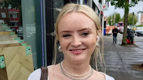 Food prices remain 'stubbornly high' in Northern Ireland as inflation rate rises A young woman is standing in the street and smiling. She has blond hair that's tied back. She's wearing a white top and necklaces. She has ear pods in.