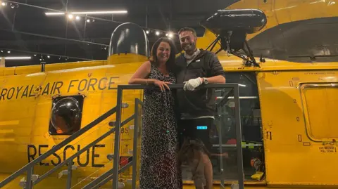 Military aircrew pursuing MoD after cancer diagnoses reaches 180 Anna-Louise Stubbings Zach standing in front of a yellow Sea King helicopter. Anna is wearing a sleeveless black and white dress and has long dark hair. Zach is in dark overalls.