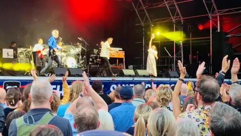 'Party city' event gets under sail Deacon Blue, a band, are on stage performing. There is a crowd clapping along.