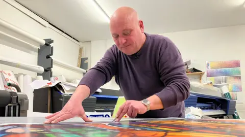 Small businesses tell of crippling costs of telecoms services BBC/Caroline Bilton A man wearing a purple jumper leaning over a table which has a multi-colour picture on it. He has a pen in his left hand which is on the picture. He is in a white room with a couple of machines in the background with rolls of paper leaning against the wall.