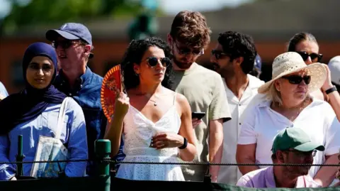 Heat continues in parts of England but temperatures cool elsewhere PA Media Spectators stand at Wimbledon - with one woman with curly dark hair and a white top standing in the middle holding a fan to cool down from the heat.