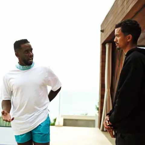 Sean Combs (left) and Phil Pines (right) talk outdoors near a building with an ocean view in the background; Combs wears a white t-shirt and blue shorts, Pines is in a black hoodie.