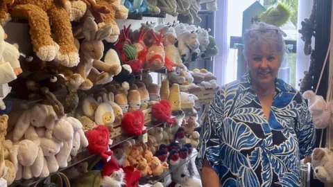 Alison McCabe A woman with short white hair, a blue and white patterned shirt, and glasses on the top of her head stands, smiling, next to a large display of Jellycat plush toys including rabbits and fish