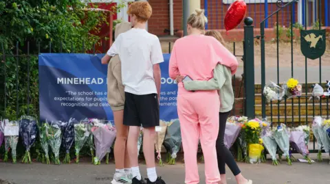 PA Media A group of people standing outside and looking at the line of floral tributes left outside Minehead Middle School. Two children are stood on the left hugging and a woman and a girl are hugging on the right. None of them are looking at the camera. 
