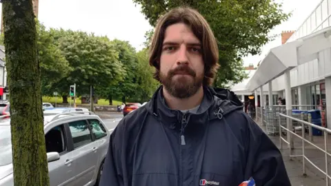 Food prices remain 'stubbornly high' in Northern Ireland as inflation rate rises A man is standing in the street outside a supermarket. He's wearing a navy jacket and has brown hair and a moustache and short beard.