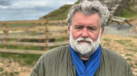 BBC Hairy Biker Si King is stood in front of a fence surrounding the stump at Sycamore Gap, wearing a blue scarf and dark green jacket on a cloudy day