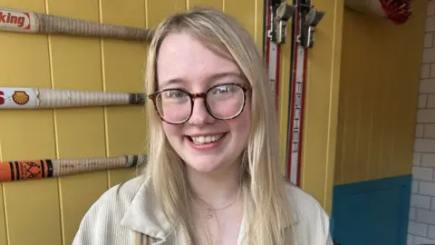 How to get one when you have zero work experience Grace Pickett A woman with long blonde hair and brown glasses in sat inside a building with yellow walls. She is smiling at the camera