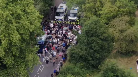 Epping council votes to urge government to shut asylum hotel Essex Police An image taken by a drone, showing police officers using vans to block a large group of people from continuing down a road. The road is flanked by tall trees.