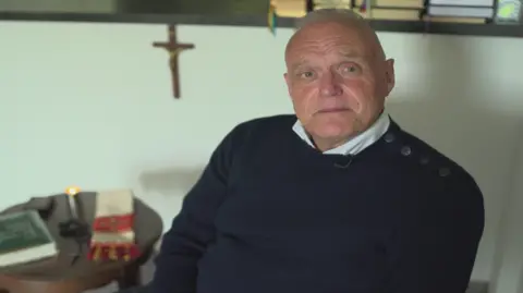 Police investigate sex abuse claims against men linked to Church A man with receding white hair is sitting down wearing a dark jumper and a white shirt in front of a cross and next to a table with books and a candle on it