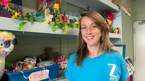 Summer holiday clubs now cost an average of £1,076 per child Elaine Doran/BBC Gilly Balfour with shoulder length brown hair wearing a blue t shirt with the logo Z arts on it smiling in front of shelves of colourful children's games and art materials