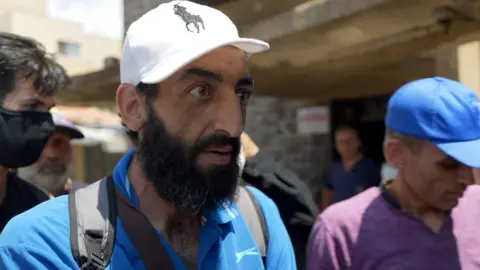'They shot patients in beds' – BBC hears claims of massacre at Suweida hospital BBC / Jon Donnison Kiness Abu Motab wears a white ralph lauren designer baseball cap, a grey backpack with straps and a blue slazenger polo shirt. He is looking away from the camera at a reporter out of shot