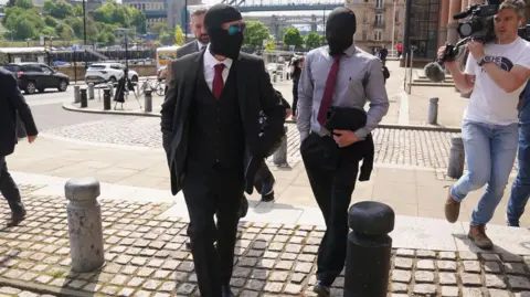 PA Daniel Graham and Adam Carruthers are walking into court but they have covered their faces with balaclavas. A man with a camera is walking alongside them. 