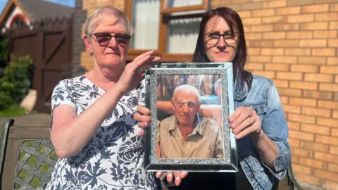'Hospital gave us two death certificates for dad to cover up their mistake' BBC Two women hold up a silver framed picture of an older man.
The woman on the left, Susan Holmes, is wearing a black and white floral patterned top and dark glasses and has short blonde hair while the woman on the left, Lisa Jones, has long dark hair and is wearing a denim jacket and glasses. The man has short white hair and is looking directly at the camera while wearing a striped shirt. Behind them you can see the wall and window of a house.