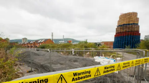 PA Media yellow tape reads WARNING ASBESTOS in front of a tall bonfire. Houses are visible in the distance