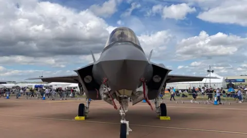 Defence firms 'confident' hundreds more jobs will come A military fighter jet, stands on a red runway. Behind it, behind fencing, crowds of people walk past and look on. There are tents behind them.