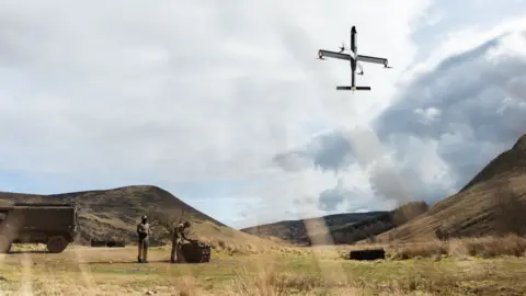 Drone manufacturer STARK to begin production in Swindon STARK Two people, bottom left, stand next to equipment, which is flying a drone that is captured in the sky to the right of the photo. They are standing in a hilly landscape with trimmed grass