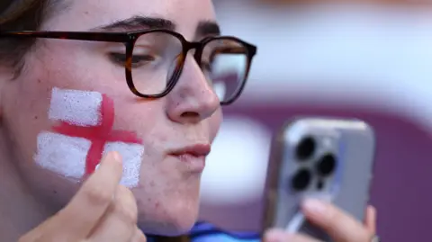 How England fans are preparing for the Euro 2025 final Reuters A woman wearing glasses is drawing a red and white England flag on her right cheek in face paint while looking at her reflection on her mobile phone.