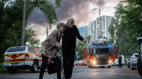 Kyiv hit by barrage of drone strikes as Putin spurns Trump's truce bid DSNS An elderly woman and man with ambulances and smoke in the sky behind them