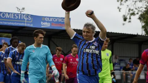 Pete Doherty plays in non-league football friendly for Margate FC Margate FC Pete Doherty raising his arms and a brown hat to the crowd as he leads out a football team in blue and black kits, which he is also wearing.