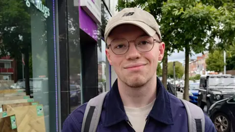 Food prices remain 'stubbornly high' in Northern Ireland as inflation rate rises A young man is wearing a cap and glasses and a navy jacket. He's standing in the street. A backpack is on his shoulders.