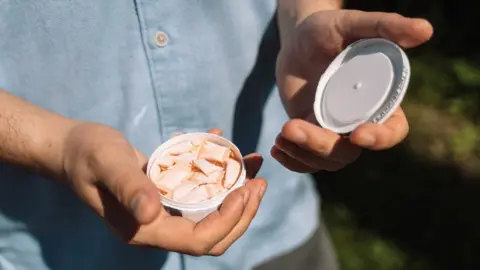 The rising use of nicotine pouches snus Getty Images A man shows the camera a small container of nicotine pouches, which look like very small pillows. He is wearing a blue button-down shirt and standing outside.