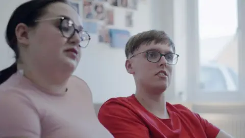 Dubious sales tactics at two leading estate agencies uncovered by BBC investigation Olivia and Ryan pictured sitting next to each other, from a slight sideways angle, on a sofa in a living room. Olivia is slightly out of focus in the foreground, she has long, dark hair tied back, and a tight-fitting pink top. She is wearing glasses. Ryan has short fair hair with a fringe, black-rimmed glasses and is wearing a red-T-shirt.