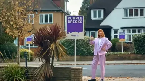 Dubious sales tactics at two leading estate agencies uncovered by BBC investigation Still taken from an advert, showing a woman standing on a suburban pavement in front of 1930s homes. There are Purplebricks for sale signs in front of three houses. She is wearing a pink suit and has her thumb up.