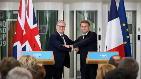 UK-France migrant deal 'robust' against legal challenges, Yvette Cooper says Reuters Sir Keir Starmer and Emmanuel Macron shake hands in front of two podium.