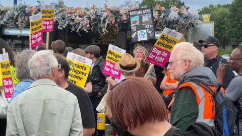 Officers attacked at Epping asylum hotel protest, police say Simon Dedman/BBC A group of people stand together with some holding banners that read "Refugees Welcome - Stop the far right". They are standing beside a building with flowers on the roof, and a green space behind it.