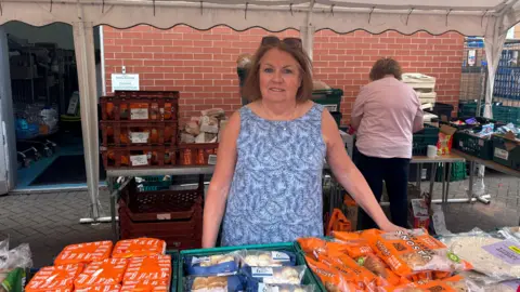 Coventry food charity user says missing meals to feed kids is the norm BBC Diane Williams, wearing a blue patterned dress and sunglasses on her head, stands in front of a table of food at the social supermarket, Grub Hub, in Coventry