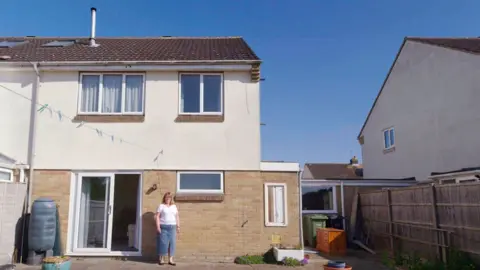 Dubious sales tactics at two leading estate agencies uncovered by BBC investigation Picture of Julie's house taken from the back garden. It is a 1980s semi-detached home with sliding patio doors. She is standing to the right hand side of the doors. It is a sunny day.