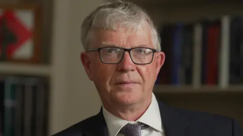 Police investigate sex abuse claims against men linked to Church Sir Malcolm wears a black suit, white shirt and patterned tie. He has short white hair and wears dark-rimmed glasses. He looks straight at the camera without smiling.