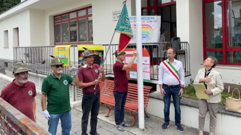 'The village will die' - Italy looks for answers to decline in number of babies School closing ceremony - men in Alpine hats hold a flag and a bugle