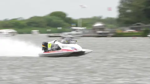 Powerboat driver dies after crash with houseboat at Oulton Broad A single white and black powerboat races from left to right with water spraying behind it. In the background are blurry buildings and trees, and flagpoles.