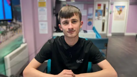 Package holidays to Spain, Cyprus and Turkey soar in price Abi Smitton / BBC News A man with brown hair sits at a desk, wearing a black polo neck shirt.