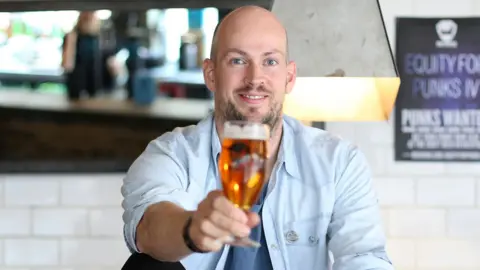 Brewdog to close 10 pubs across UK, some 'within days' Getty Images A smiling bald, bearded man wearing a light blue shirt - with the sleeves rolled up - and a light blue t-shirt raises a full beer glass with his right hand. He has been photographed in a pub setting, with a white wall and a bar visible in the blurry background.