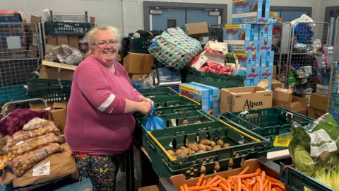 Coventry food charity user says missing meals to feed kids is the norm A smiling Alainea Stark wearing a dark pink jumper and leggings stands in a store room next to a table with crates of vegetables including carrots and potatoes, with other food goods stacked behind her