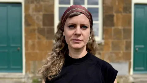 Investigating one of the UK's most abusive cults Ellena Wood with long curly hair, a head scarf and a black top stands in front of a small orange-brick chapel building.
